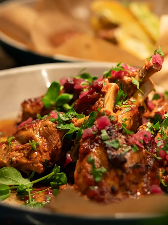 Saftige lammekoteletter serveres i en skål, pyntet med friske urter og terninger av rødløk. Tallerkener med annen mat, som salater, er uklare i bakgrunnen, noe som antyder en måltidssituasjon.