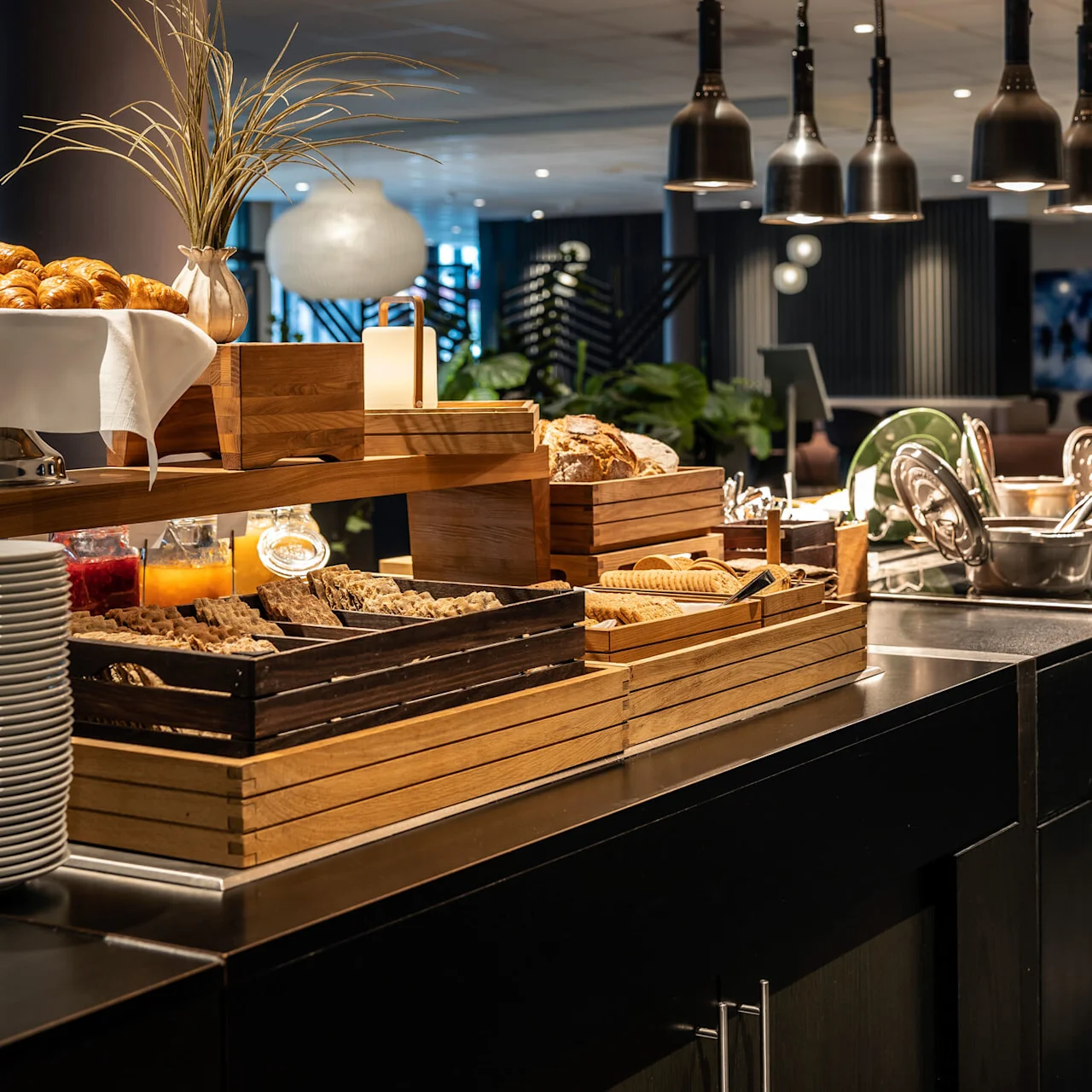 En buffet med croissanter, brød, kornprodukter og bestik i trækasser. Overliggende lamper oplyser madopstillingen med grønne planter og siddepladser i baggrunden.