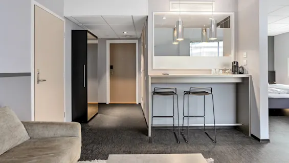 A modern hotel room with an open plan featuring a kitchenette, couch, and a glimpse of a bed, showcasing a clean and minimalist aesthetic.