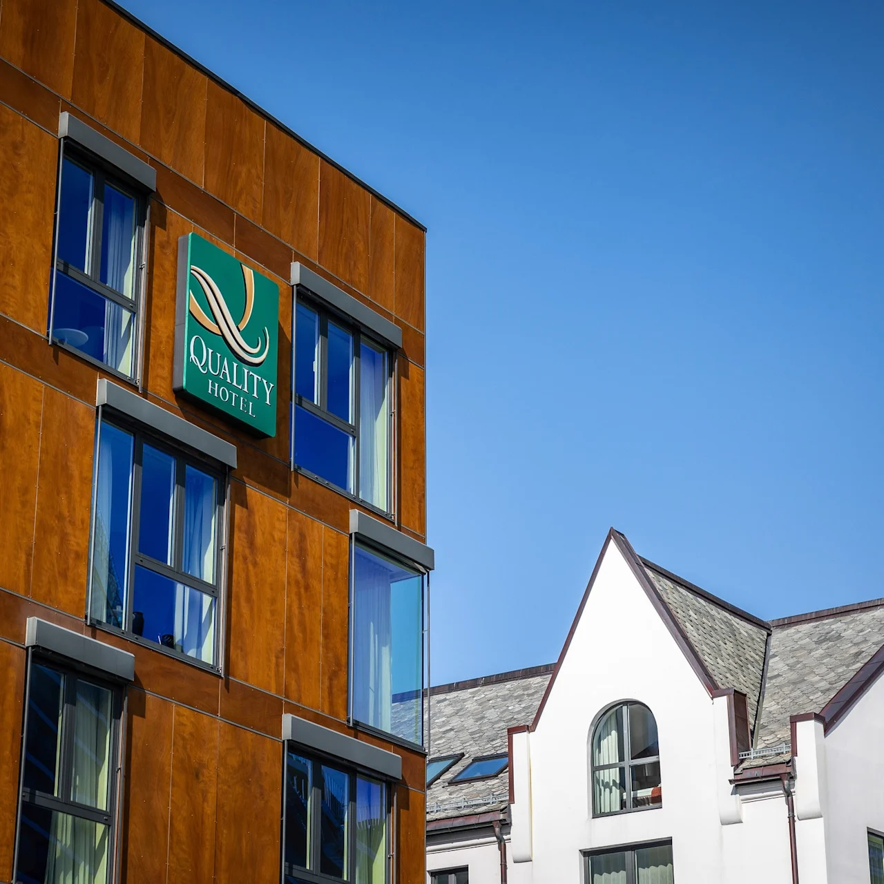Fasaden på en brun byggnad har en skylt med texten "QUALITY HOTEL" mot grön bakgrund; intill finns en vit byggnad med grått tak under en klarblå himmel.