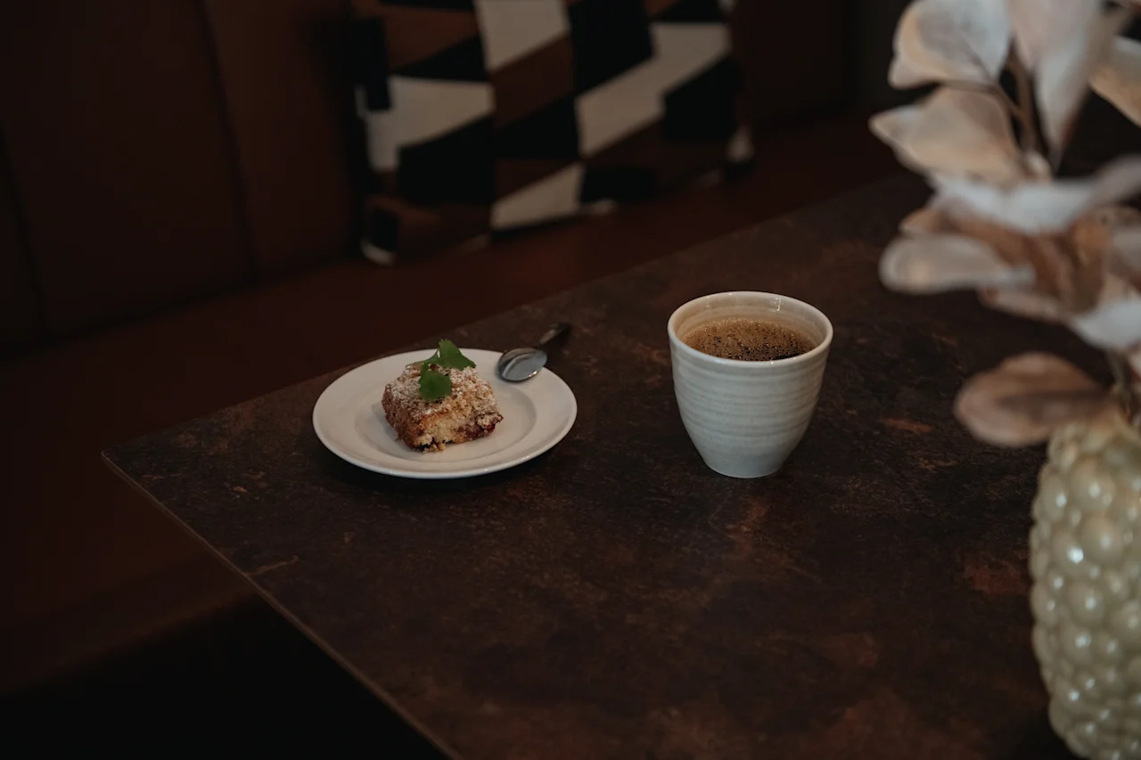 En deilig kakestykke med en skje på en hvit tallerken ved siden av en kopp kaffe på et mørkt trebord.