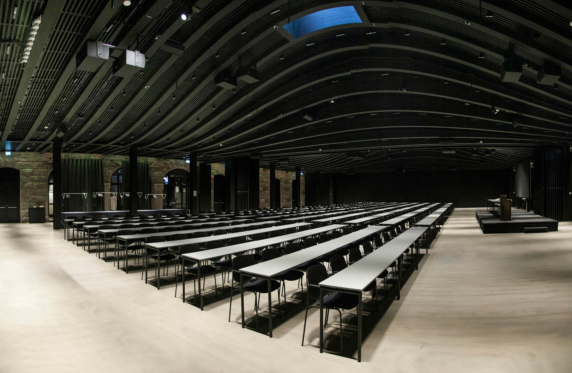 Långa bord med stolar är arrangerade i rader mot en podium i ett rymligt, mörkt takbelagt konferensrum. Väggarna är kantade med fönster och gardiner under omgivningsbelysning.