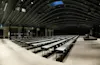 Long tables with chairs are arranged in rows facing a podium in a spacious, dark-ceilinged conference room. The walls are lined with windows and curtains under ambient lighting.