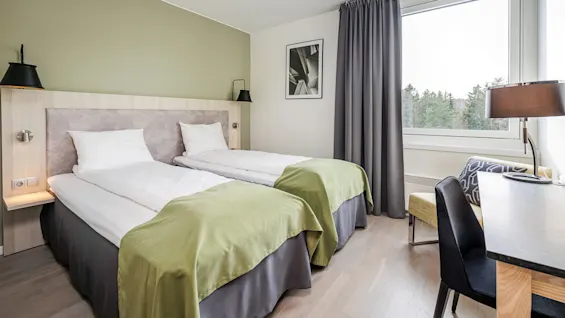 Two neatly-made twin beds with green accents, flanked by modern lamps, in a bright room with wooden flooring, a desk, and a forest view through a large window.