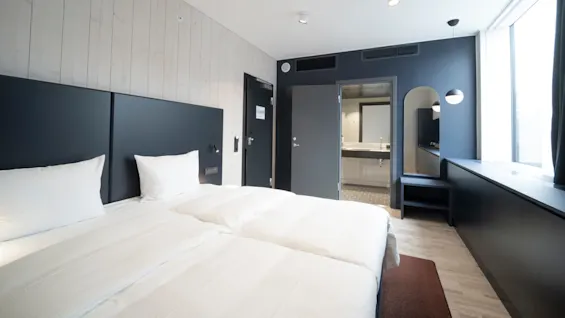 Bed positioned against a modern hotel room wall, flanked by a door leading to a bright bathroom featuring a large mirror, with a window illuminating the relaxed ambiance.