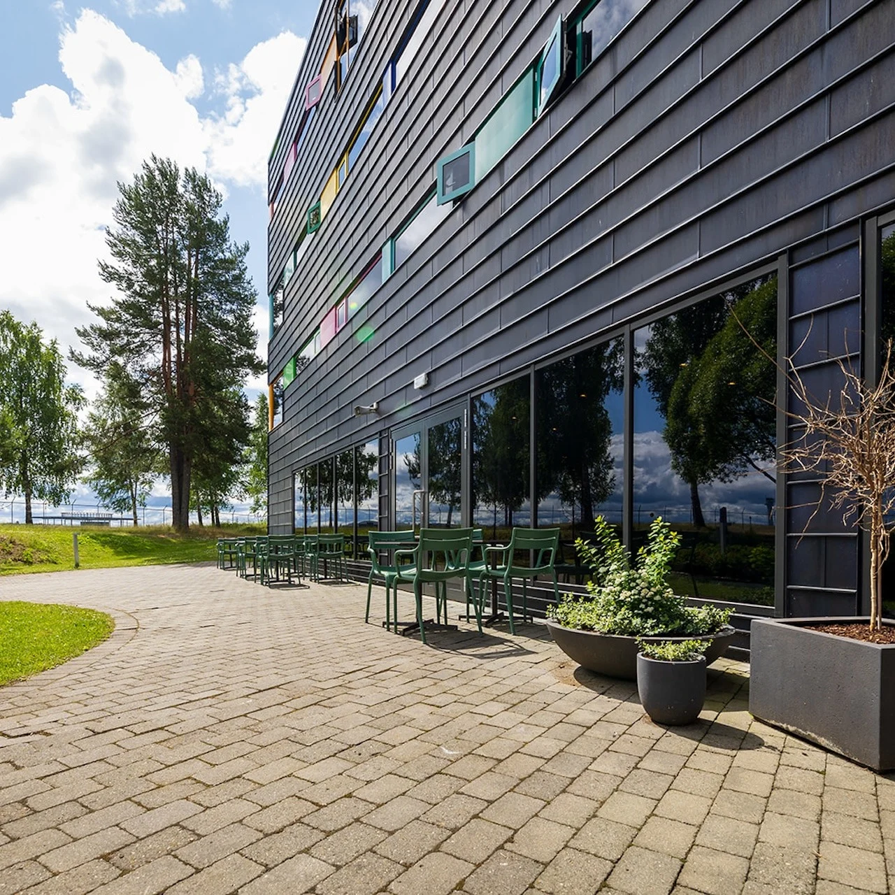 Building with reflective glass windows and green chairs on a paved patio extends into a grassy area with trees under a cloudy sky.