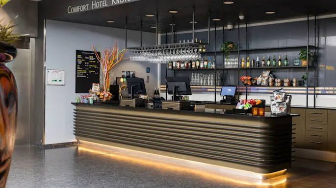 A modern hotel reception desk displays snacks and drinks under hanging glasses in a well-lit lobby. Above, the sign reads "COMFORT HOTEL KRISTIANSAND."