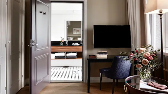 Døren åbner til et badeværelse med håndklæder pænt stablet på hylder; et mørkt skrivebord med bøger og blomster er i nærheden, ved siden af gardiner og en lampe.