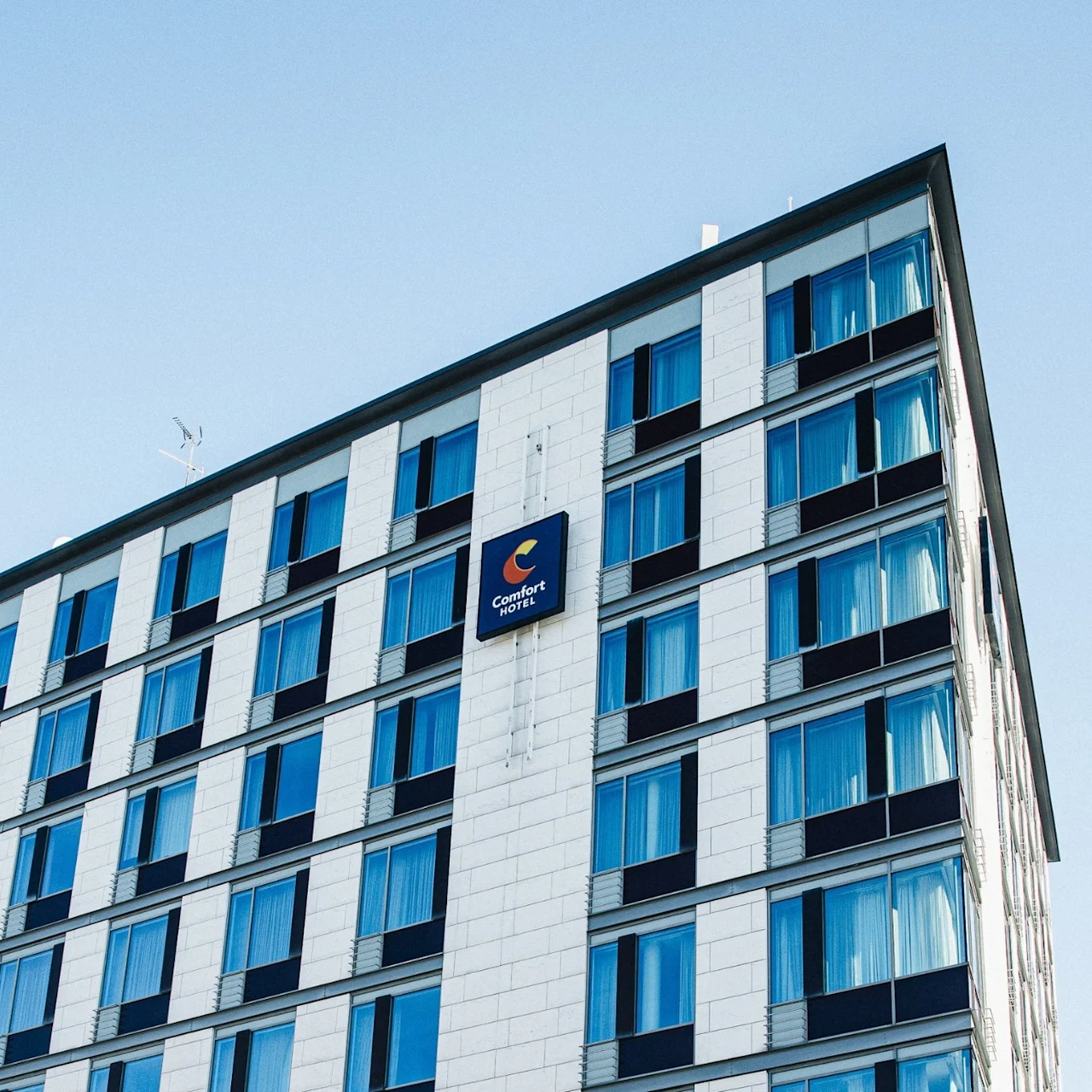 A multistory building stands with a sign reading "Comfort Hotel," featuring blue-tinted windows, set against a clear sky.