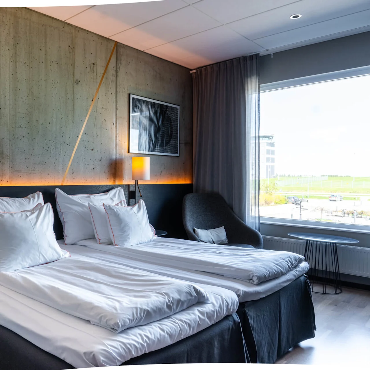 En prydligt bäddad säng med vita lakan och kuddar bredvid en stilren lampa, placerad i ett modernt, välbelyst hotellrum med stora fönster och utsikt utanför.