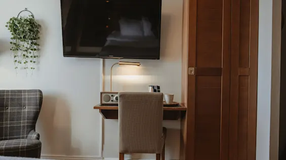 Et hyggeligt hotelværelse med et tv monteret over et skrivebord, en lampe og en behagelig stol. En plante hænger på væggen.
