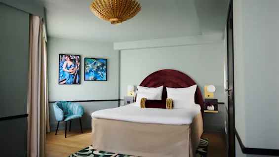 Bed sits centered, adorned with red headboard and white bedding, framed by side tables and lamps. Context includes patterned rug, artwork on pale walls, and a teal chair.