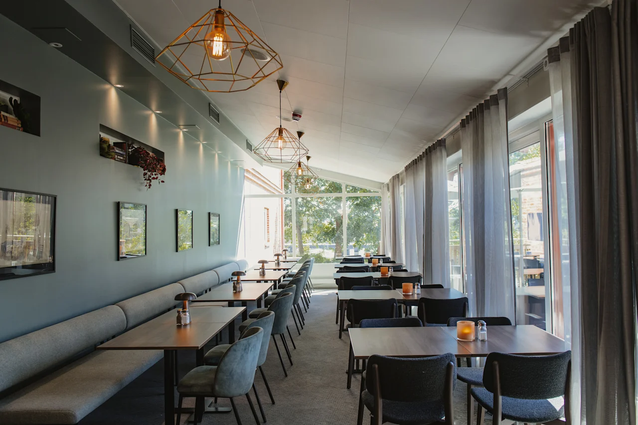 En ljus och modern matplats med bord, stolar och bänkplatser, upplyst av naturligt ljus och geometriska taklampor.