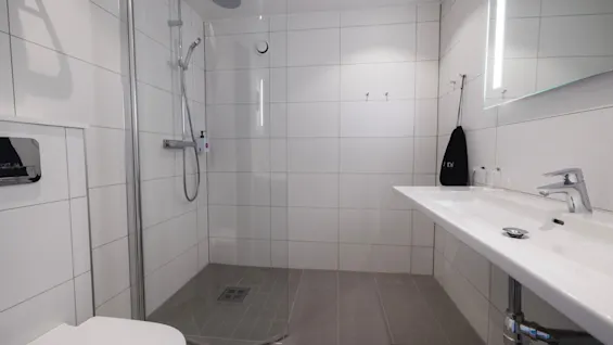 A modern bathroom features a glass-enclosed shower, wall-mounted toilet, and wide sink with a mirror. The walls are tiled in white, and the floor has dark tiles.
