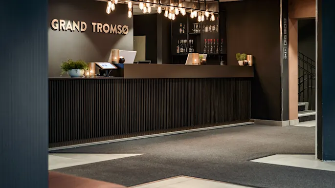 A modern hotel reception area with a dark wood desk, illuminated by stylish hanging lights. Text on the wall reads GRAND TROMSO.