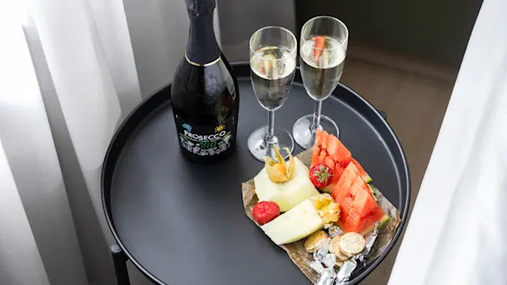 Et lite svart bord har en flaske musserende og to glass, med et fargerikt utvalg av skivet frukt og sjokolade, som skaper en innbydende setting for en intim feiring ved vinduet.