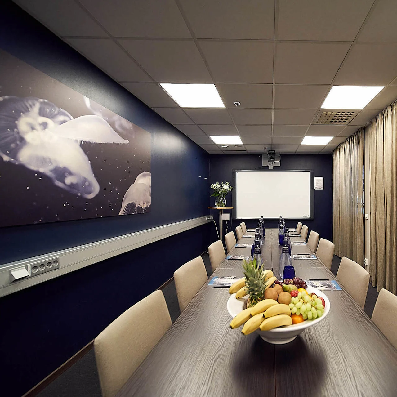 Et langt konferansebord med fruktskål i midten, omgitt av stoler i et svakt opplyst rom med mørkeblå vegger, en manetmaleri, beige gardiner, og en hvit projektorskjerm.