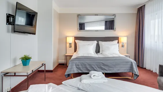 A tidy hotel room with a double bed, mounted TV, and large window draped with sheer and opaque curtains.