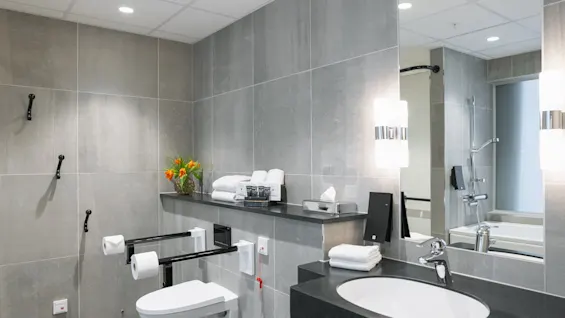 Modern badrum med grå kakel, handfat med spegel, toalett med handtag, nya handdukar, orange blommor i vas och dämpad belysning, vilket skapar en ren atmosfär.