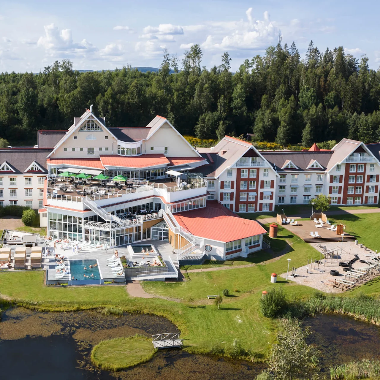 Et stort hotell med røde tak og hvite vegger har gjester som nyter et utendørs svømmebasseng og solbading. Omgitt av frodig grønn skog med en terrasse med sitteplasser.