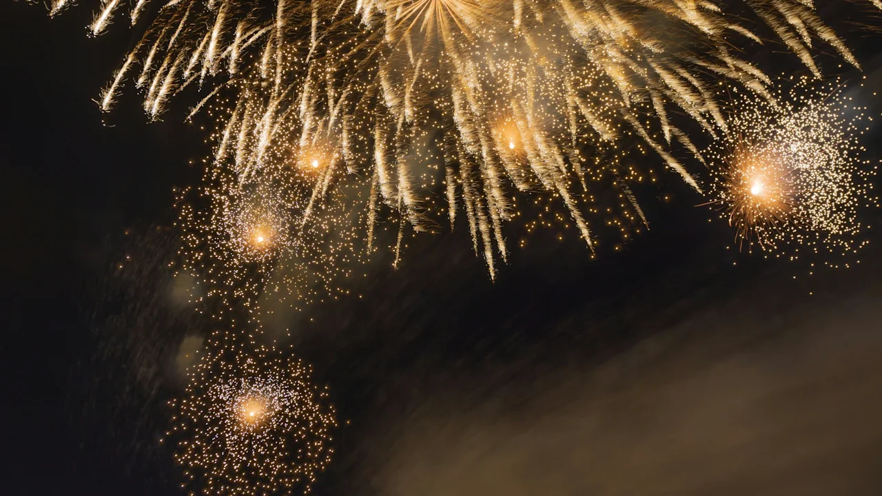 Fireworks explode in a night sky, illuminating the dark backdrop with golden light. A crowd of onlookers observes the spectacle, capturing the moment with cameras and phones.