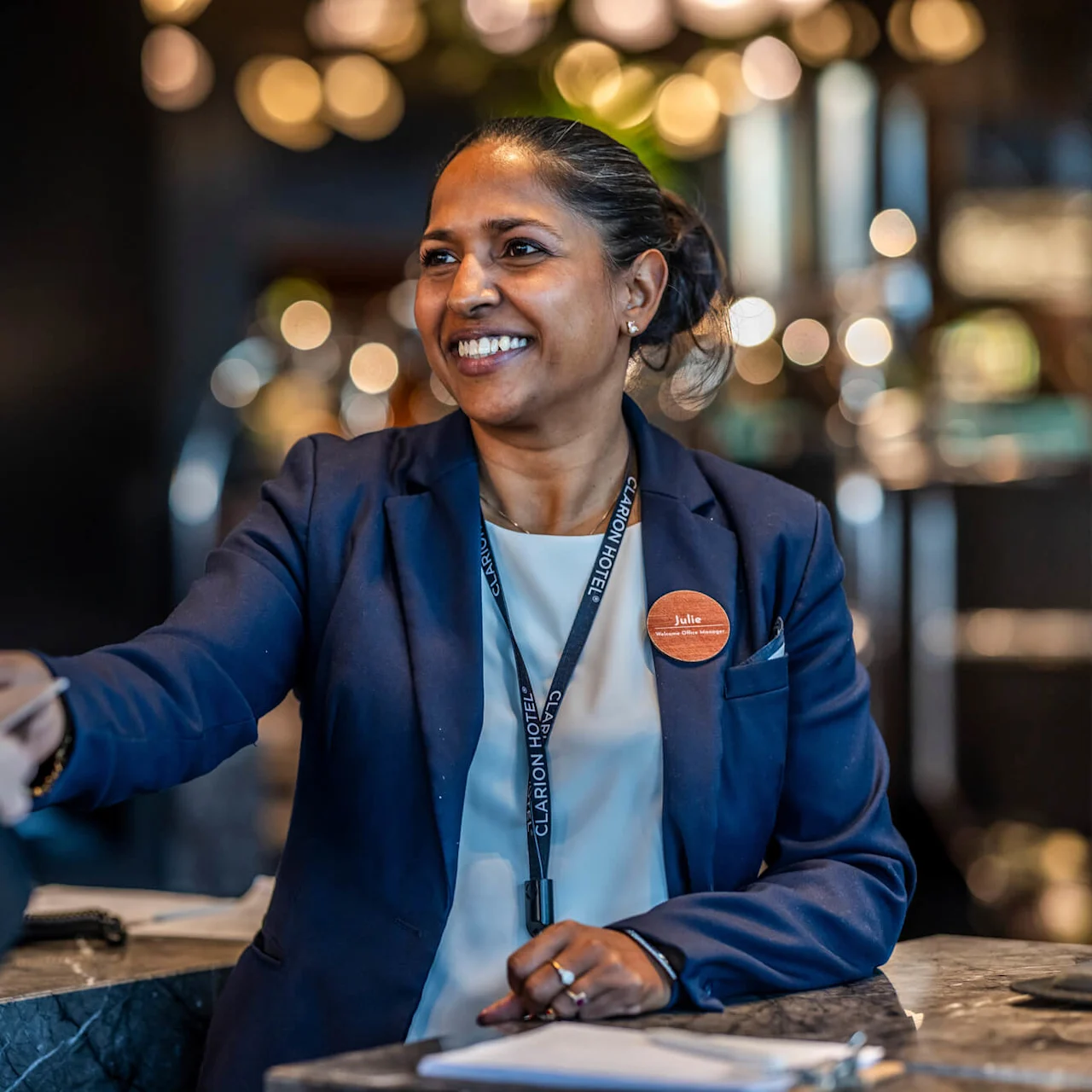 En receptionist med en "Julie"-namnbricka och Clarion Hotel-nyckelband ler medan hon hjärtligt skakar hand med en gäst över disken i en dovt upplyst hotellobby.