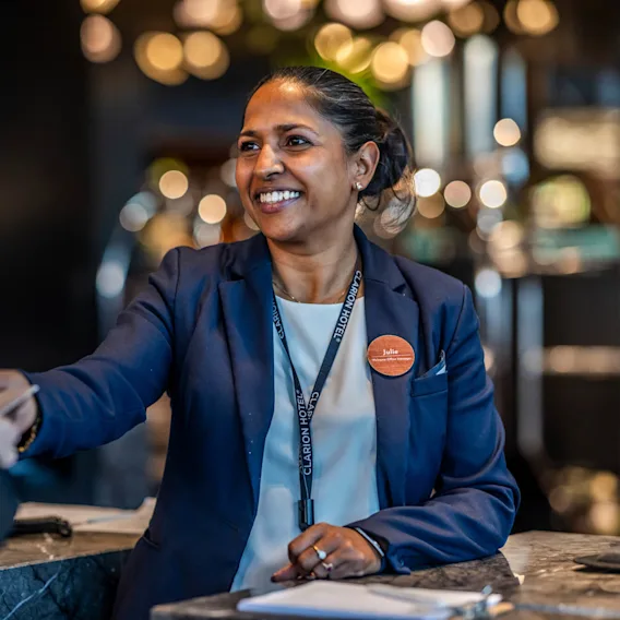 En receptionist med navneskilt "Julie" og Clarion Hotel-nøglebånd smiler, mens hun varmt giver en gæst hånden over skranken i en blødt oplyst hotellobby.