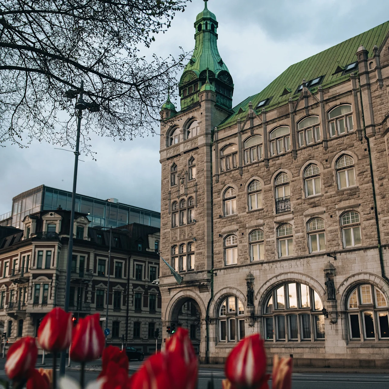 A historic building with arched windows and a green roof stands surrounded by city architecture and blooming red tulips in the foreground. The word "SPARBANK" is engraved on it.