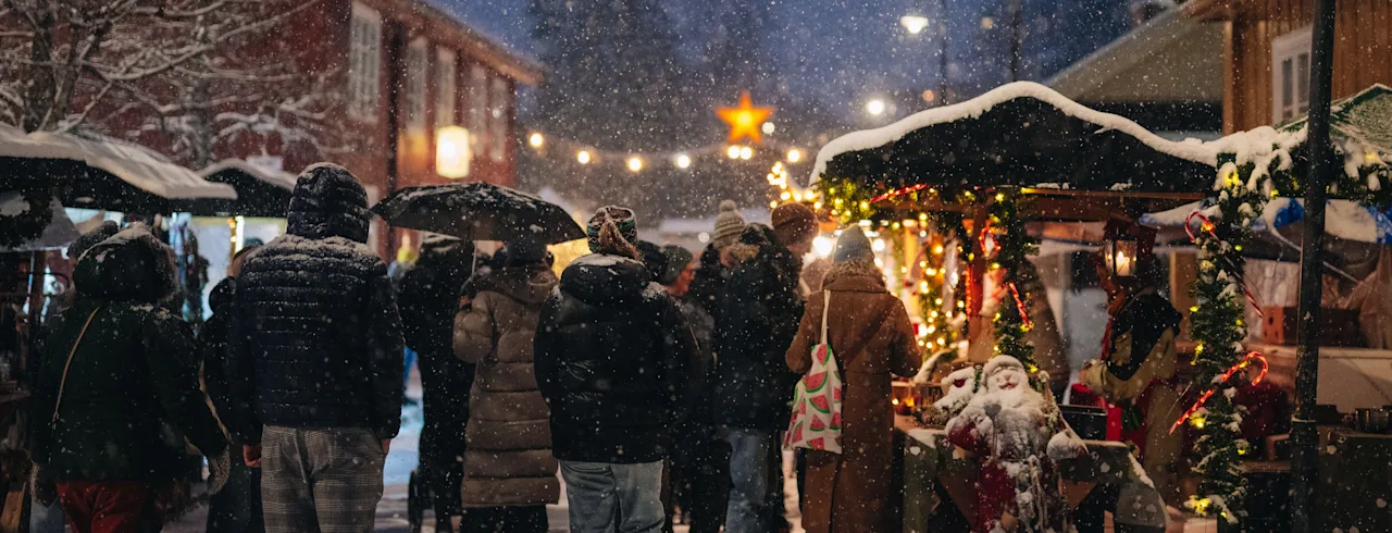 People strolling through a cozy outdoor Christmas market in the falling snow under warm festive lights