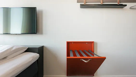 En väggmonterad, röd bagagehylla är utfälld i ett hotellrum bredvid en säng och under en TV. En trähylla med galgar är ovanför.