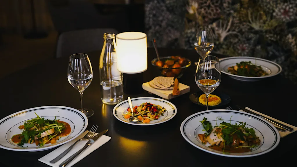 Et svakt opplyst spisebord viser tallerkener med gourmetmat, inkludert løvverk og fargerike garnityrer, omgitt av vinglass, en vannflaske, en liten lampe og diverse snacks.