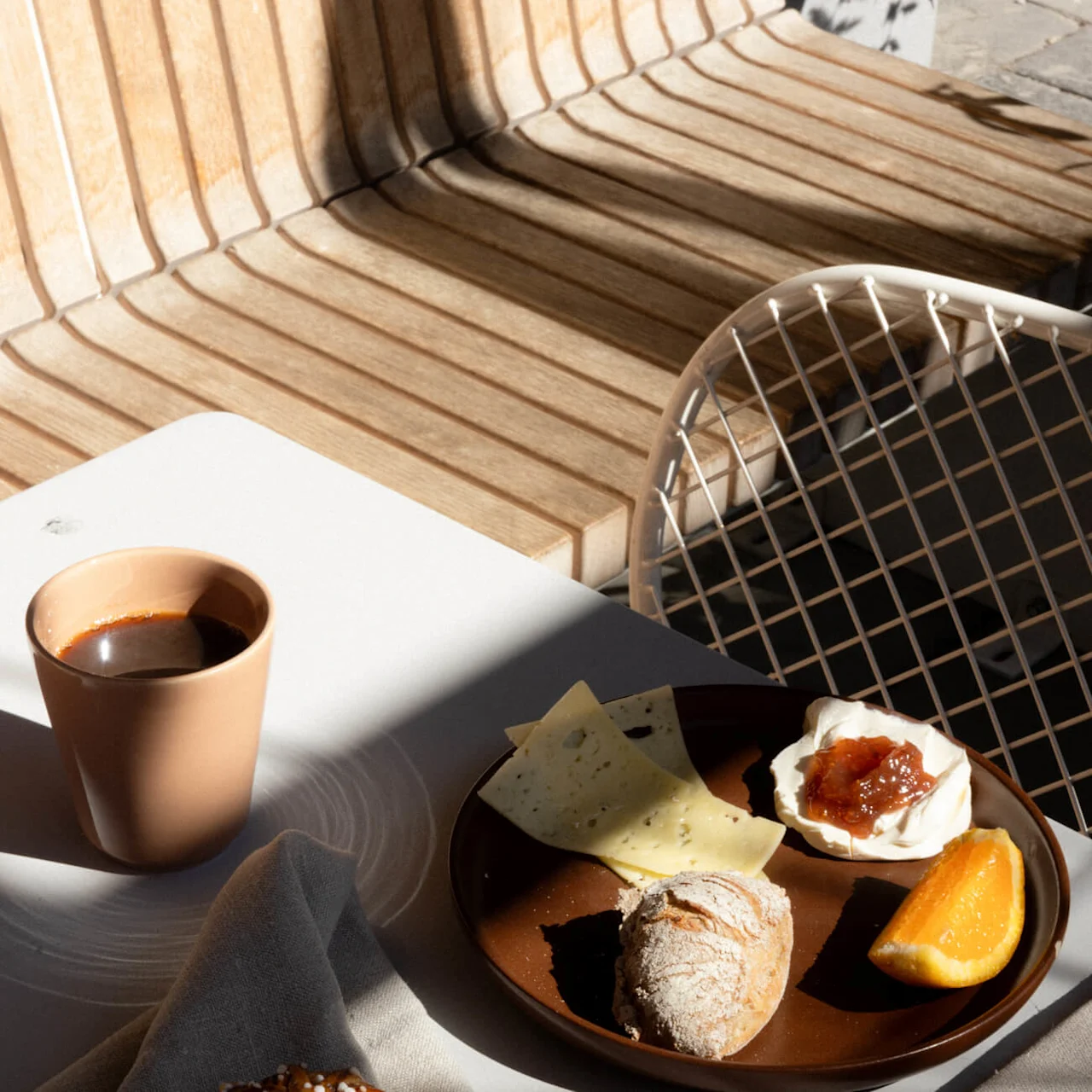 Breakfast setting with pastry sitting on a napkin, coffee cup, plate with cheese, jam, bread, orange slice, and a small glass of orange juice on a white table. Sunlit wooden bench and metal chair in the background.
