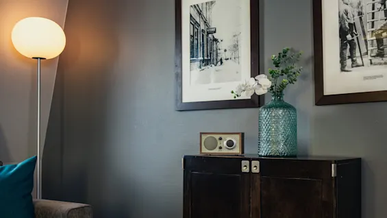 Ett mörkt träskåp håller en grön glasvas med blommor och en liten radio. Det står mot en grå vägg med två svartvita inramade fotografier, bredvid en lysande golvlampa.