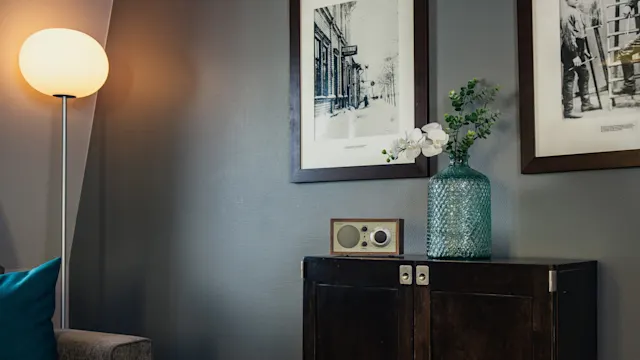 A dark wooden cabinet holds a green glass vase with flowers and a small radio. It's placed against a gray wall with two black-and-white framed photographs, beside a glowing floor lamp.