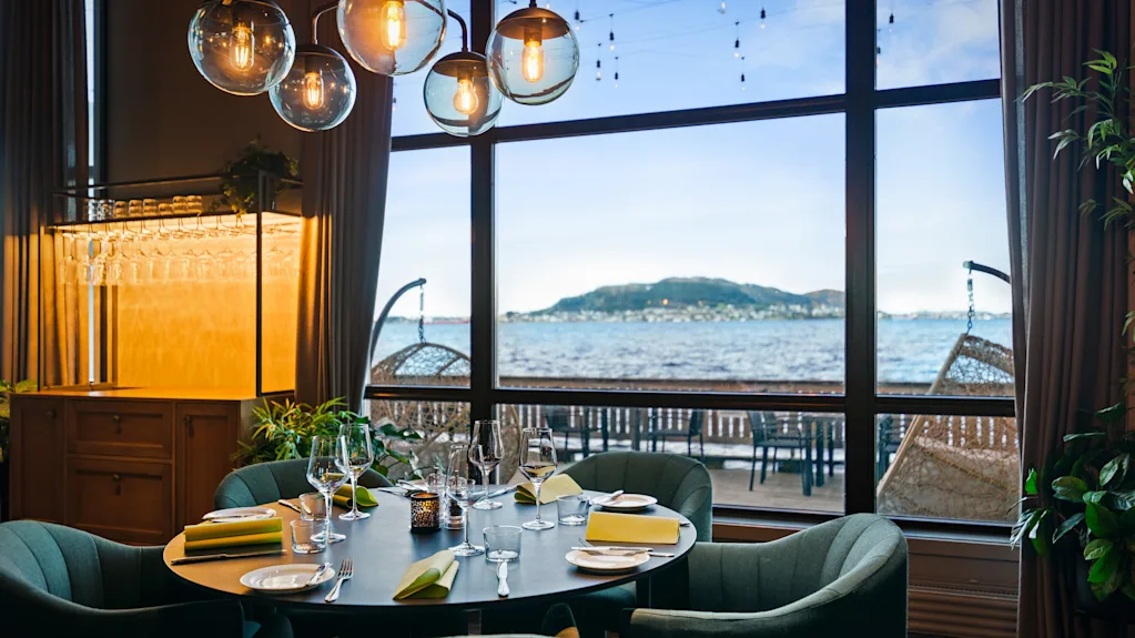 Spisebord er elegant dækket med tallerkener og glas. Moderne sfæriske lys hænger over. Stort vindue giver udsigt over kystlandskab og blå himmel med ø, som forbedrer den naturskønne spisesituation.