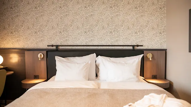 En pent oppredd seng med hvite puter og beige teppe mot en tapetsert vegg, i et koselig hotellrom med senge lamper og en stol.