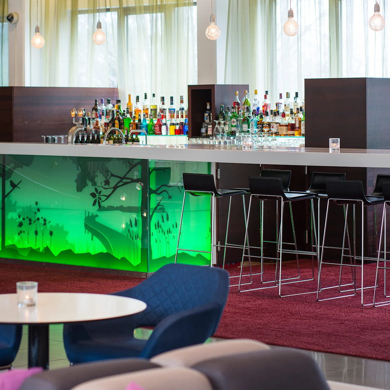 En moderne bar har en glassdisk med ulike brennevinsflasker, bakgrunnsbelyst av grønt lys, omgitt av høye stoler, i en salong med rød teppe og myke sitteplasser.