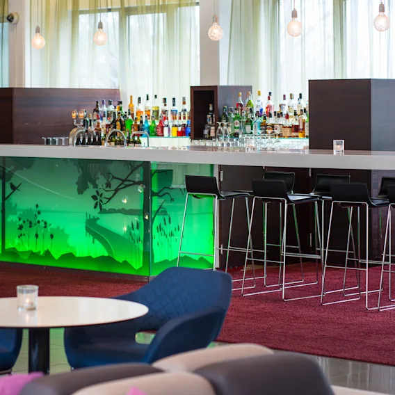 En modern bar har en glastopp med olika spritflaskor, bakgrundsbelysta av grönt ljus, omgiven av höga stolar, i en lounge med röd matta och mjuka sittplatser.