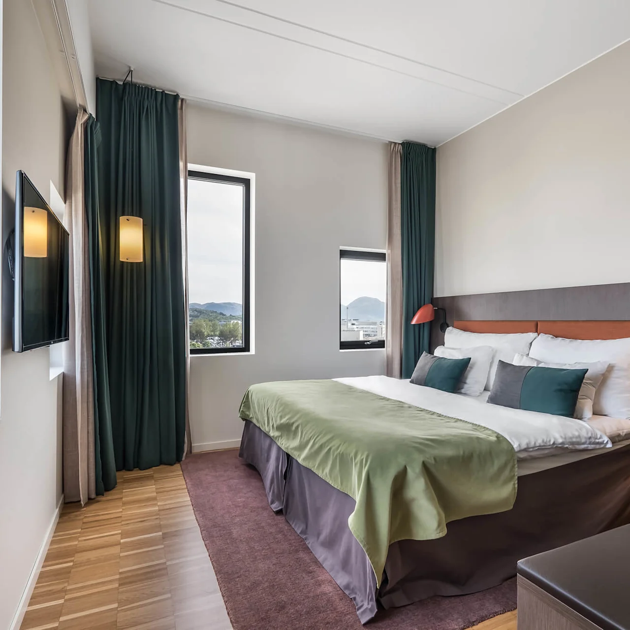 A neatly made bed with green cover stands in a modern hotel room with large windows, dark curtains, wooden floor, and a wall-mounted TV.