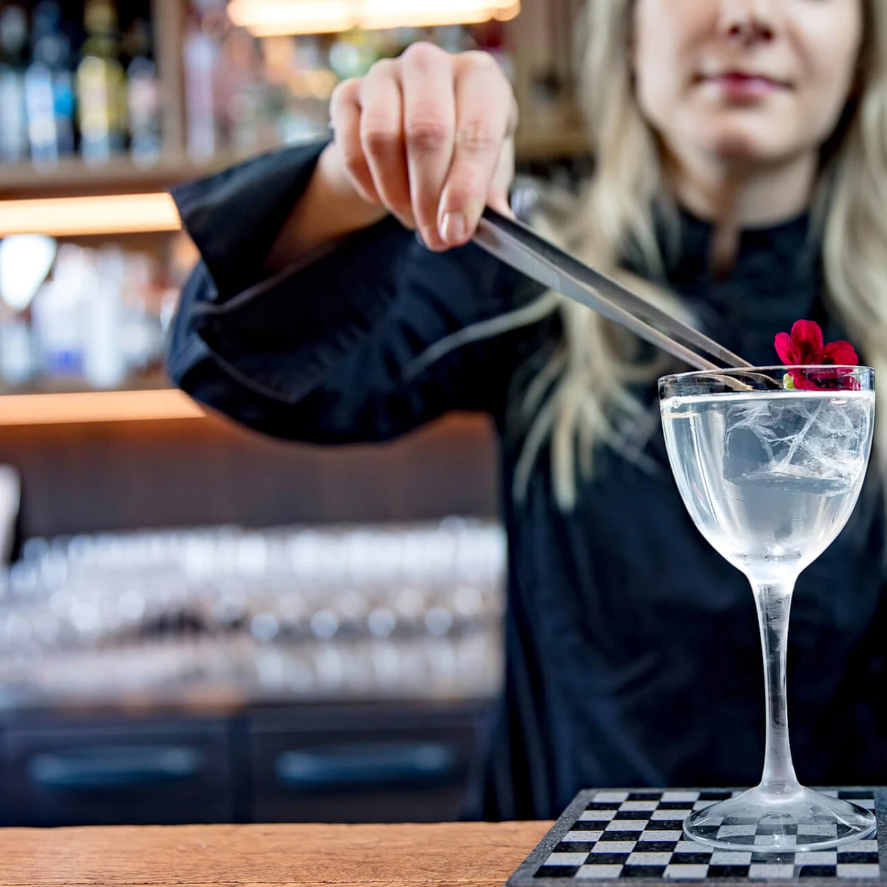 En person plasserer en rød blomst på et cocktailglass med pinsett i en bar, med flasker i bakgrunnen.