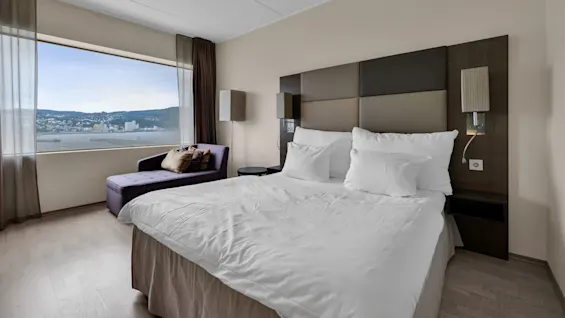 A neatly made bed with white linens is positioned beside a large window, offering a view of a bay and distant hilly cityscape. A purple chaise lounge and side table are nearby.