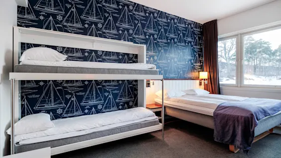 Bunk beds against a nautical-themed wallpaper depicting sailboats, beside a double bed with white linens; a large window shows a snowy landscape outside.