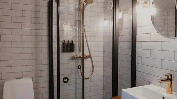 Et moderne badeværelse med bruser, toilet og håndvask. Bruseren har guldfarvede armaturer og toiletartikler. Hvide fliser pryder væggene.