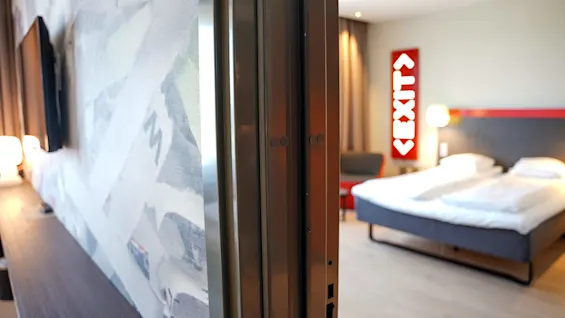 En delvis åpen dør avslører et moderne hotellrom med en stor seng og et belyst 'EXIT'-skilt.