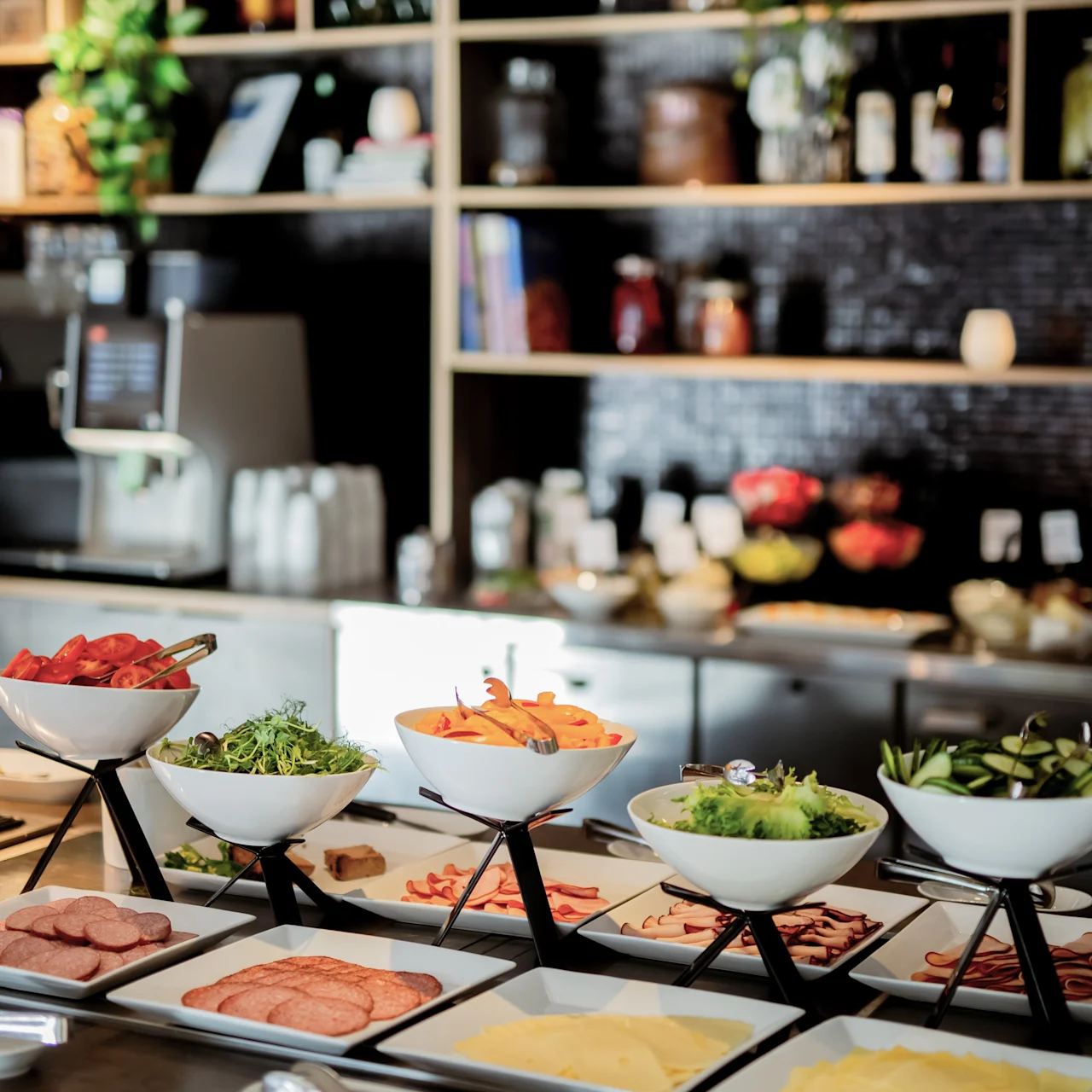 Bowls filled with vegetables and sliced meats arranged on a buffet table in a modern dining environment, surrounded by shelves stocked with jars and bottles. 