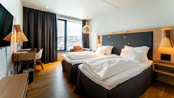 To velredte senge i et moderne hotelværelse, indrammet af trædetaljer og varm belysning, med et stort vindue viser et snedækket bylandskab.