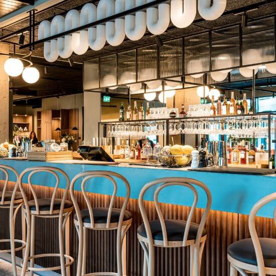 A bar with stools in a modern space, featuring hanging lights and a well-stocked counter with a bartender present.