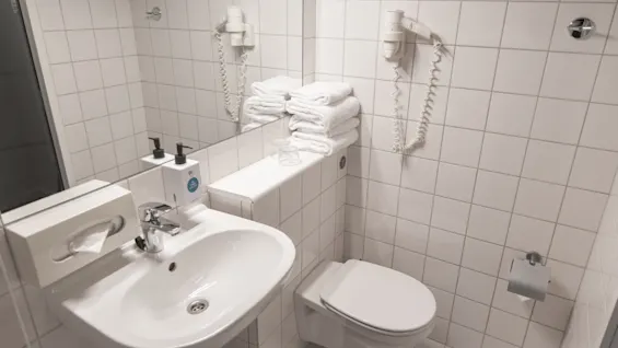 A small white-tiled bathroom contains a sink with soap dispensers, a wall-mounted toilet, a hairdryer, and a stack of towels on a shelf. A mirror reflects the scene.