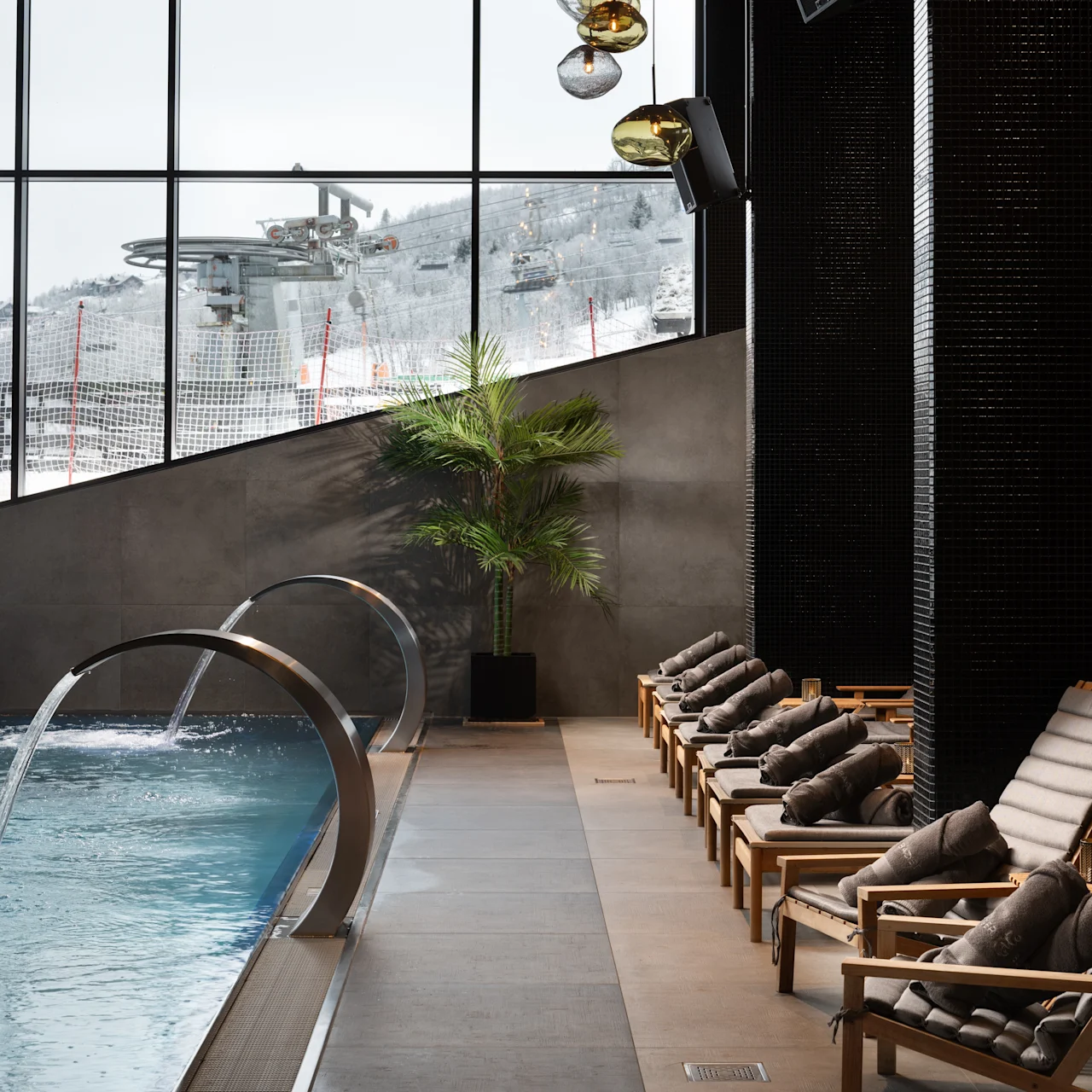 En række polstrede liggestole står langs en pool i et spa. Store vinduer viser et snedækket bjerglandskab, hvilket skaber en afslappet atmosfære.