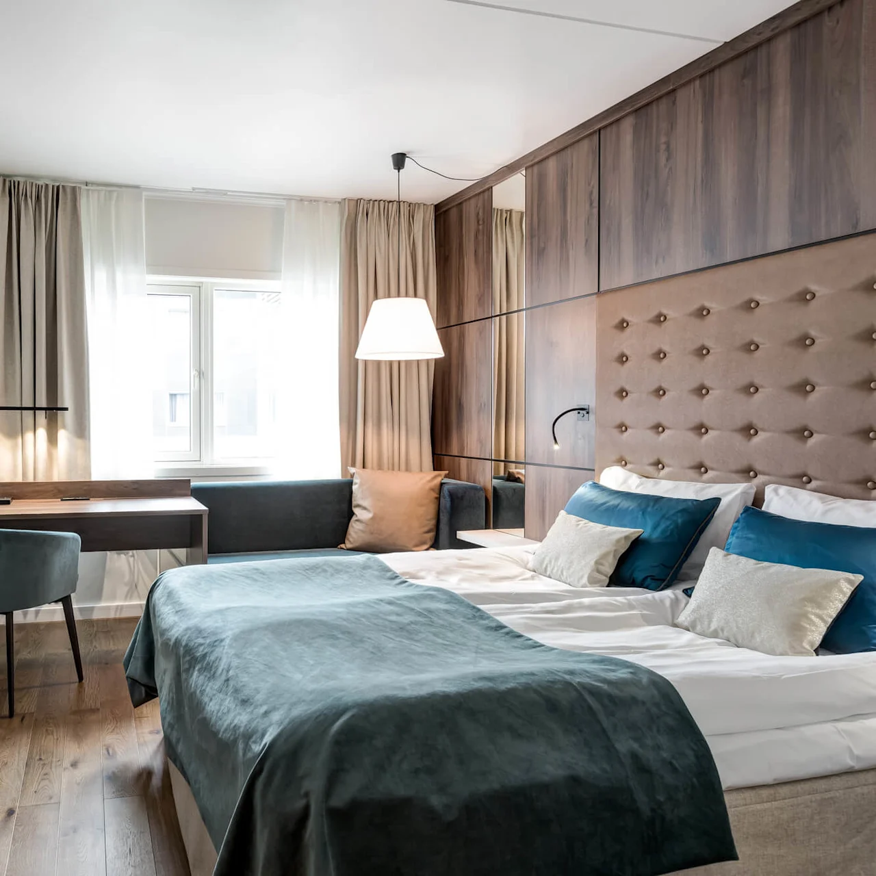 A neatly made bed with teal and white linens rests in a modern hotel room, featuring a wall-mounted TV and a desk, illuminated by natural light from the window.